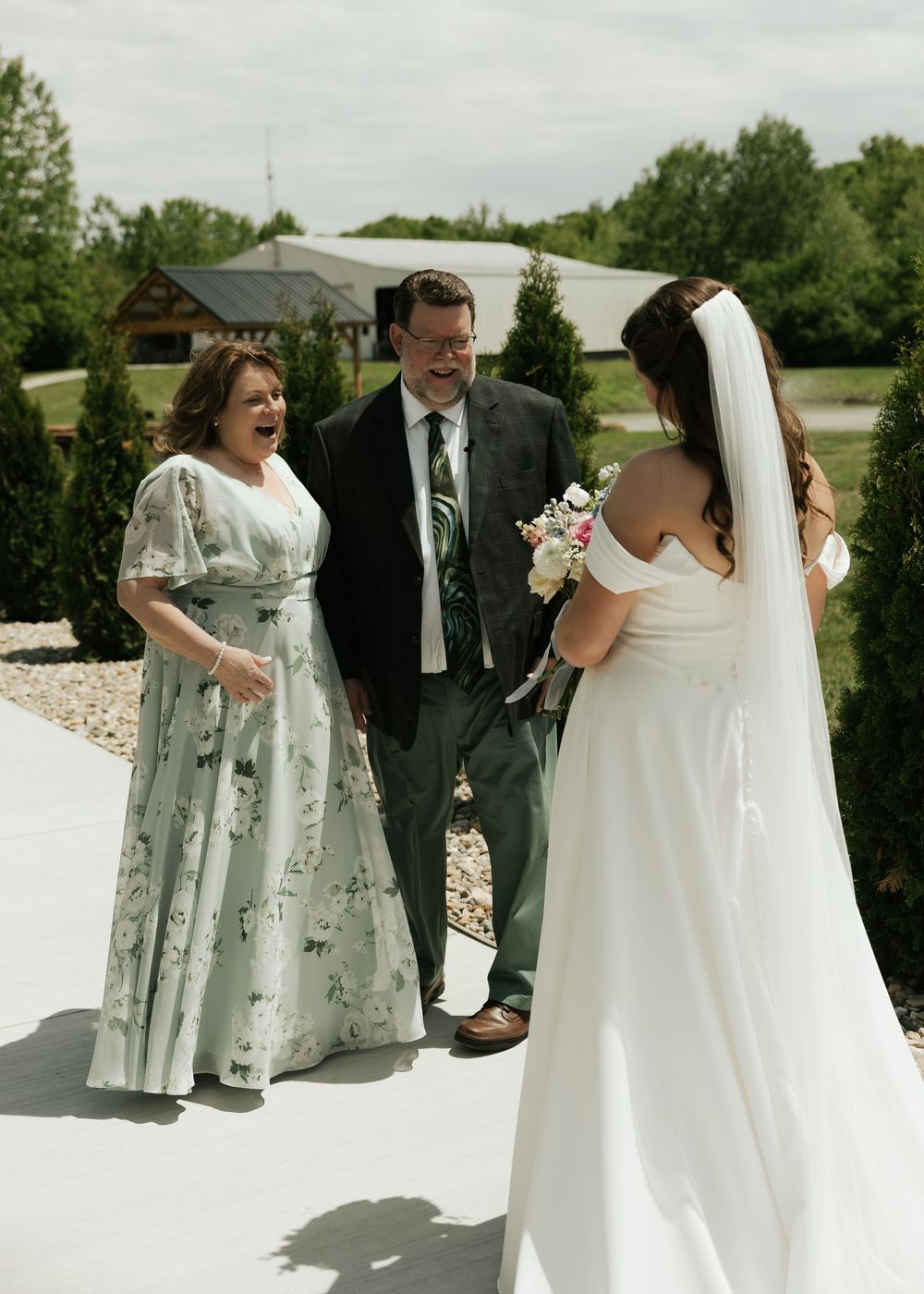 Bride in off-shoulder gown surprises parents outdoors during an emotional first look reveal on her wedding day.