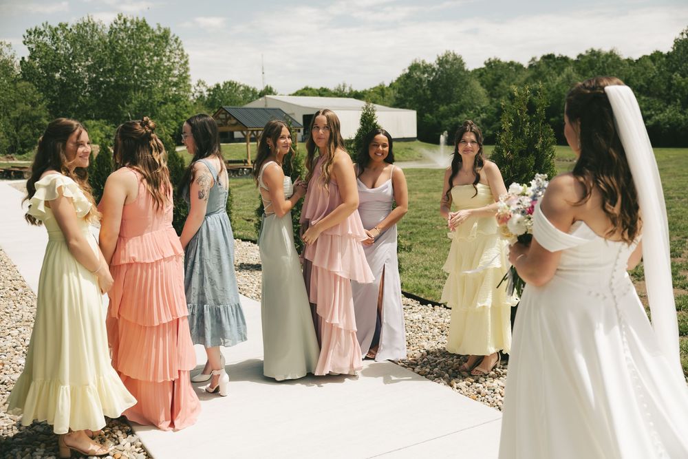 Bride faces her bridesmaids in mismatched pastel tiered dresses during a joyful outdoor first look reveal moment.