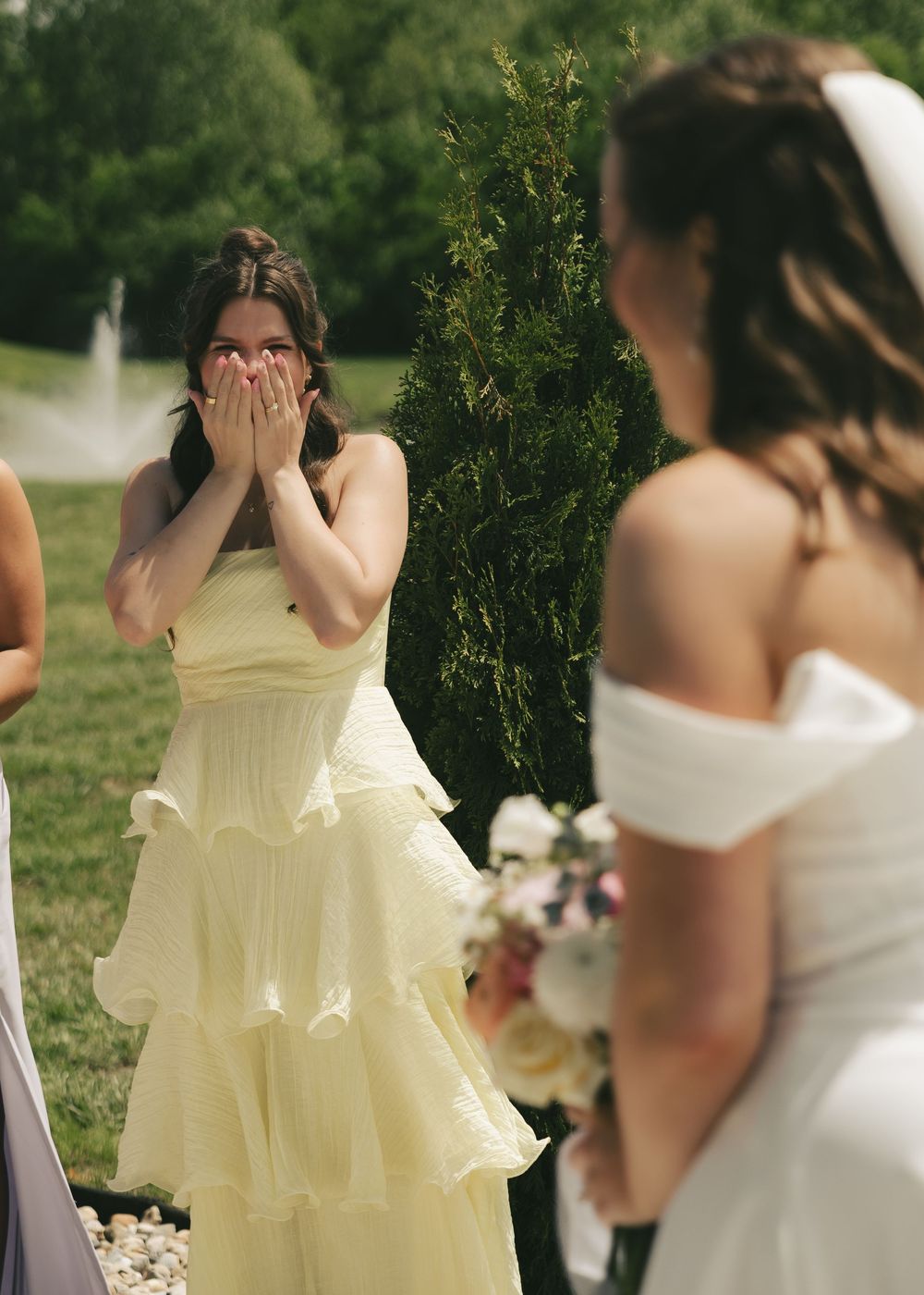 Bridesmaid in yellow ruffled dress covers her mouth in emotional reaction upon seeing the bride for the first time.
