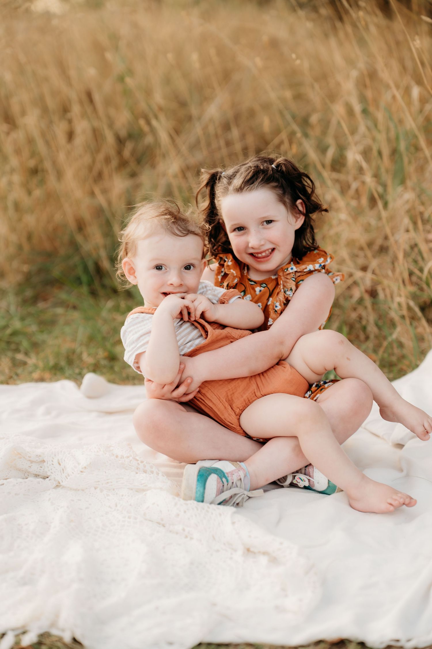 Two young siblings sit on a white blanket in a golden field, older girl hugging toddler boy, both wearing rust-toned outfits.