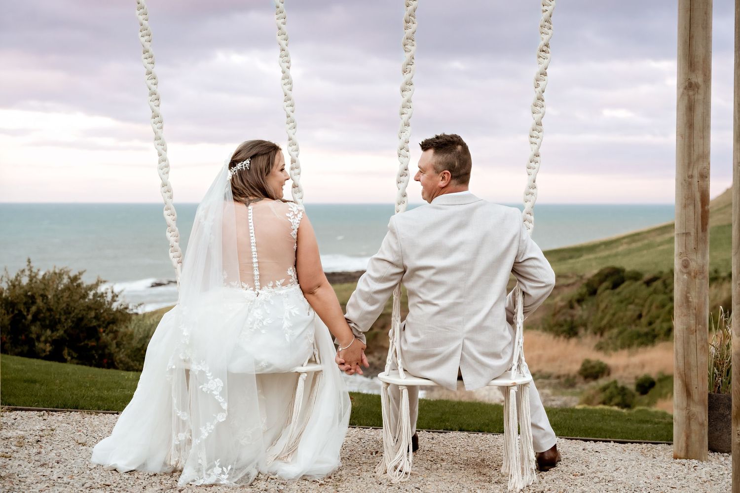 Bride and groom holding hands on a macrame swing overlooking a scenic coastal view at their outdoor wedding.