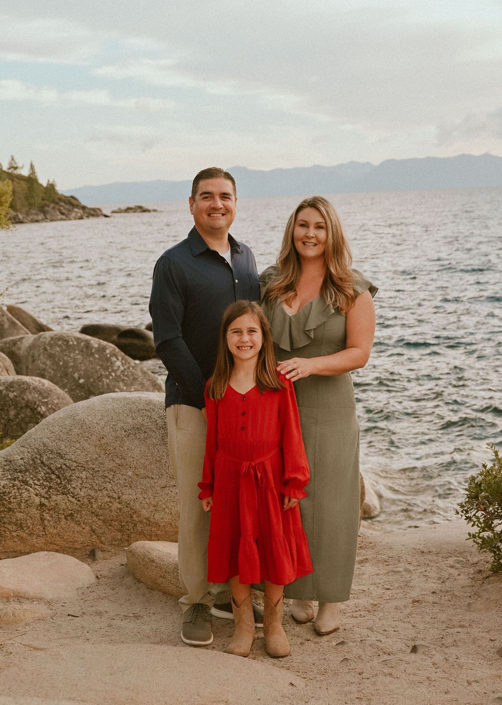Family of three posing by rocky Lake Tahoe shore, man in navy shirt, woman in olive dress, girl in red dress.