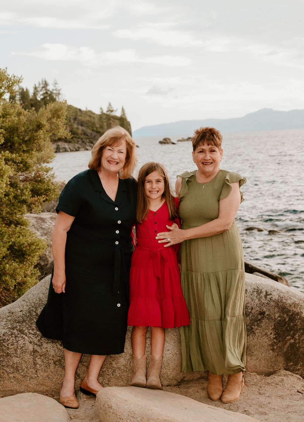 Three generations of women smiling outdoors by a lake with mountains in the background on a cloudy day.