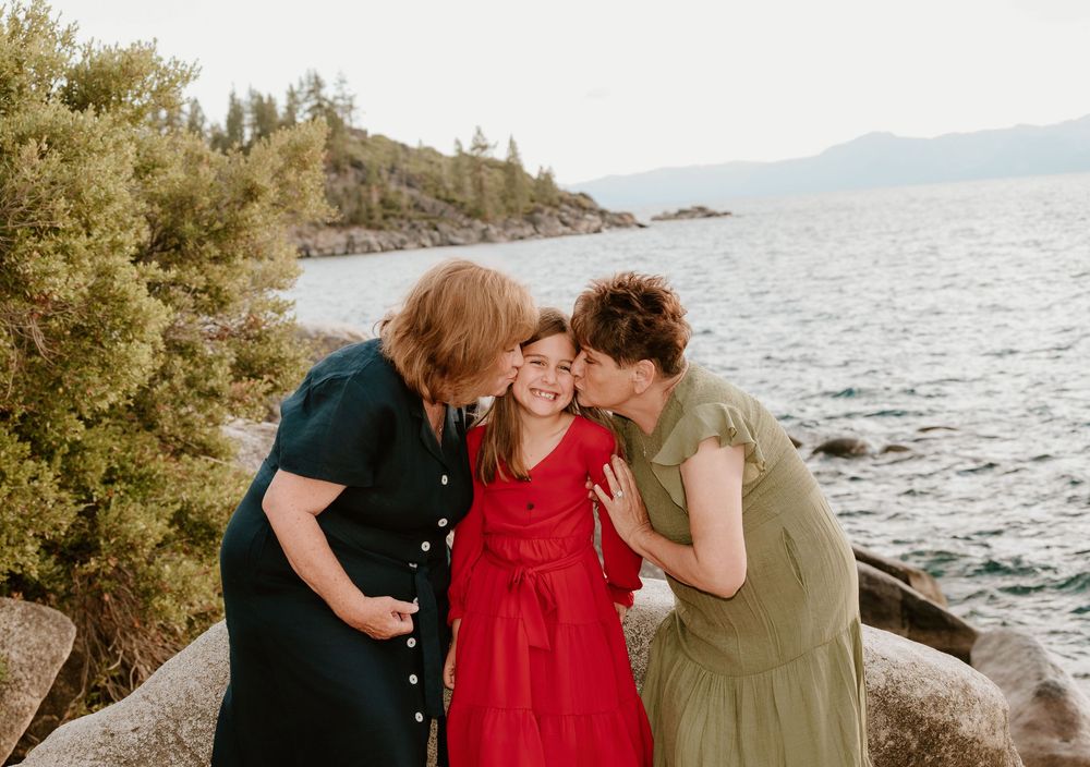 Two women kissing a young girl on the cheeks by a lake shore with rocky beach and mountain backdrop.