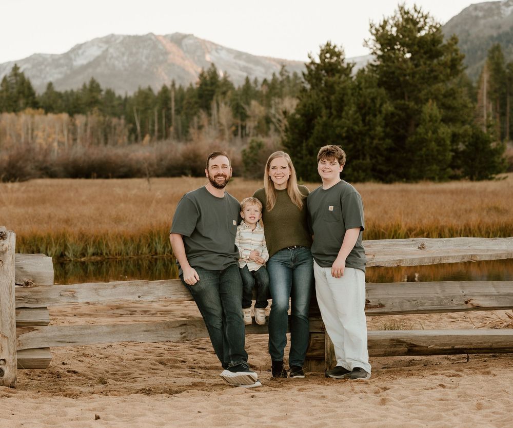 A smiling family of four poses together on sandy ground with mountains and golden fields behind them.