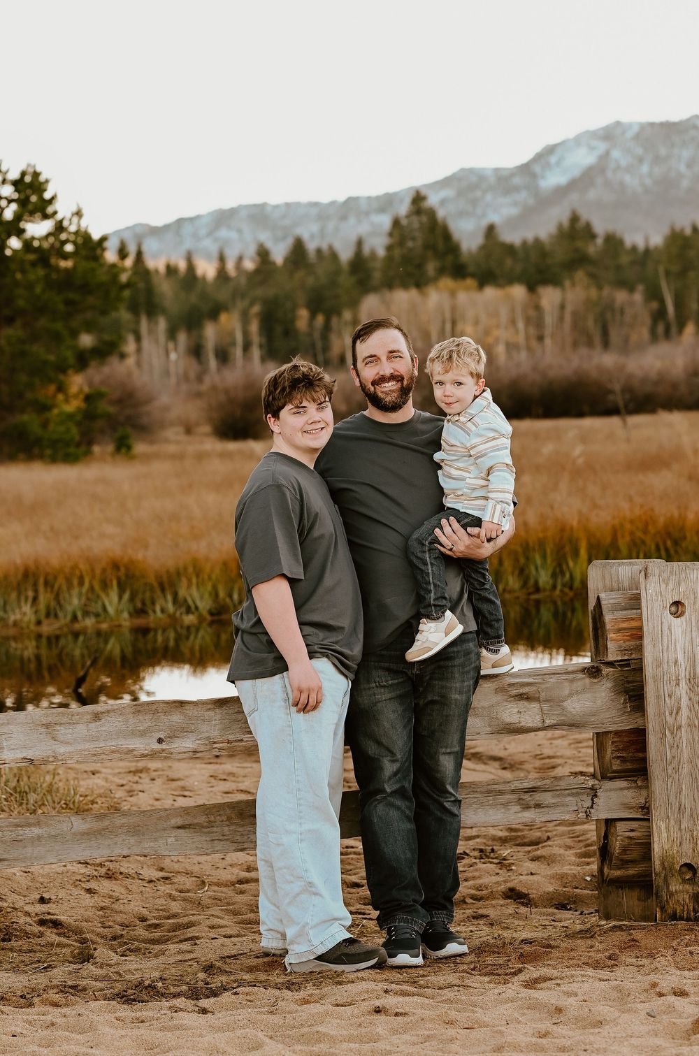 A bearded father holds a toddler while posing with his older child outdoors near a mountain lake.