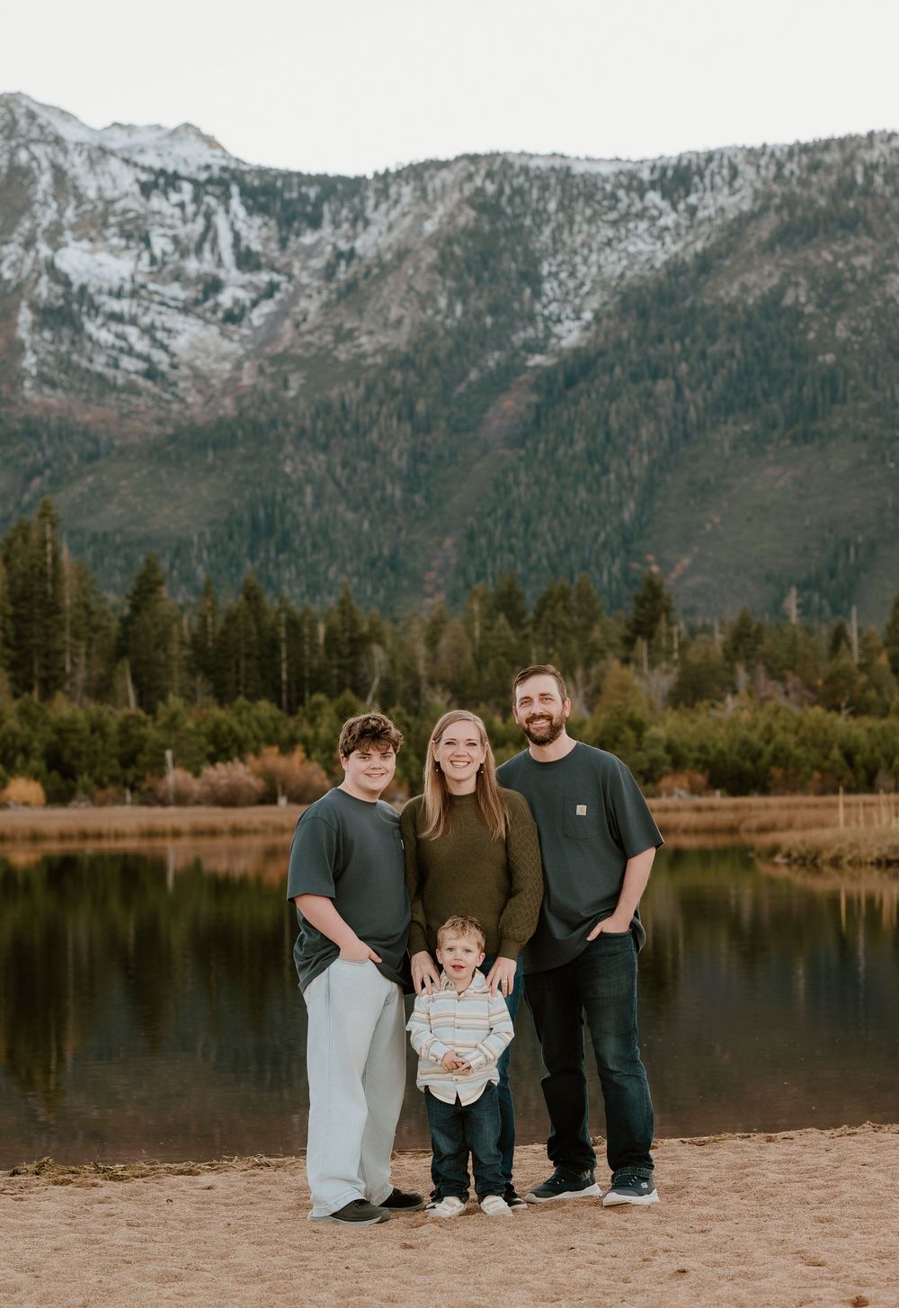 Family of four posing by a mountain lake with snow-capped peaks and pine trees in the background.