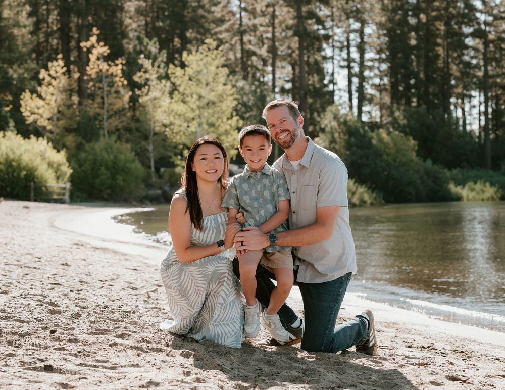 Family of three smiling on sandy beach by river surrounded by pine trees on sunny day.