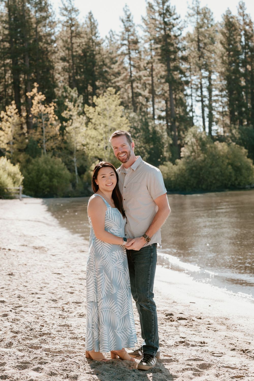 Couple embracing on sandy riverbank surrounded by tall pine trees on a sunny day.