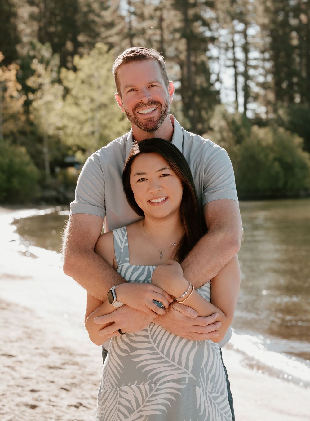 Couple embracing on a sunny lakeside beach surrounded by tall pine trees, smiling warmly in casual summer attire.