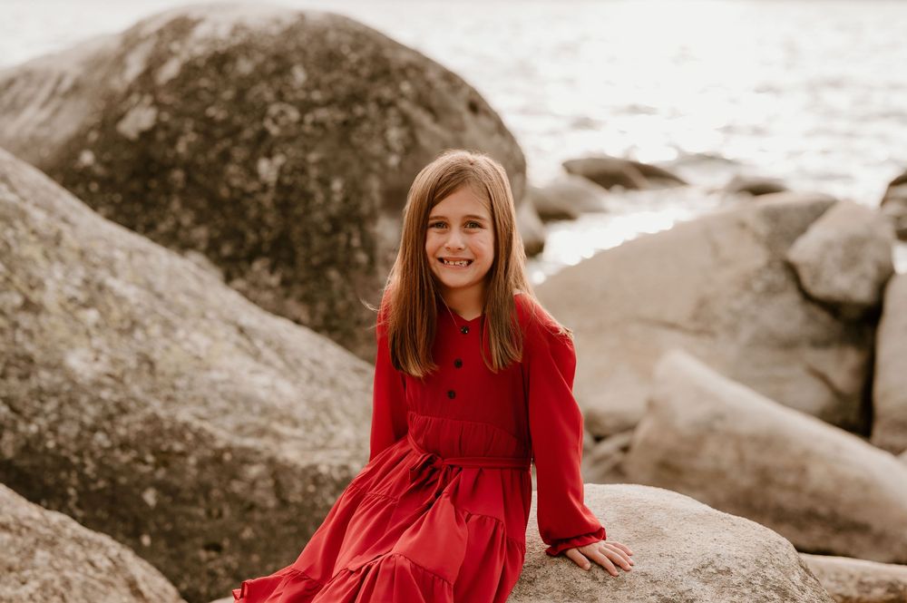 A smiling girl in a red dress sits on rocky shore with ocean in background.