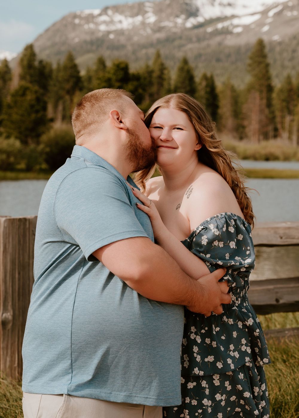 Couple sharing a romantic kiss outdoors near a mountain lake with pine trees and snow-capped peaks in the background.