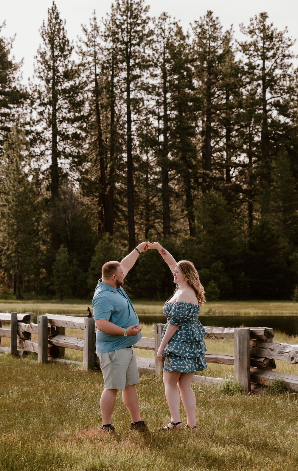 Couple dancing outdoors in a green field with wooden fence and tall pine trees in the background.