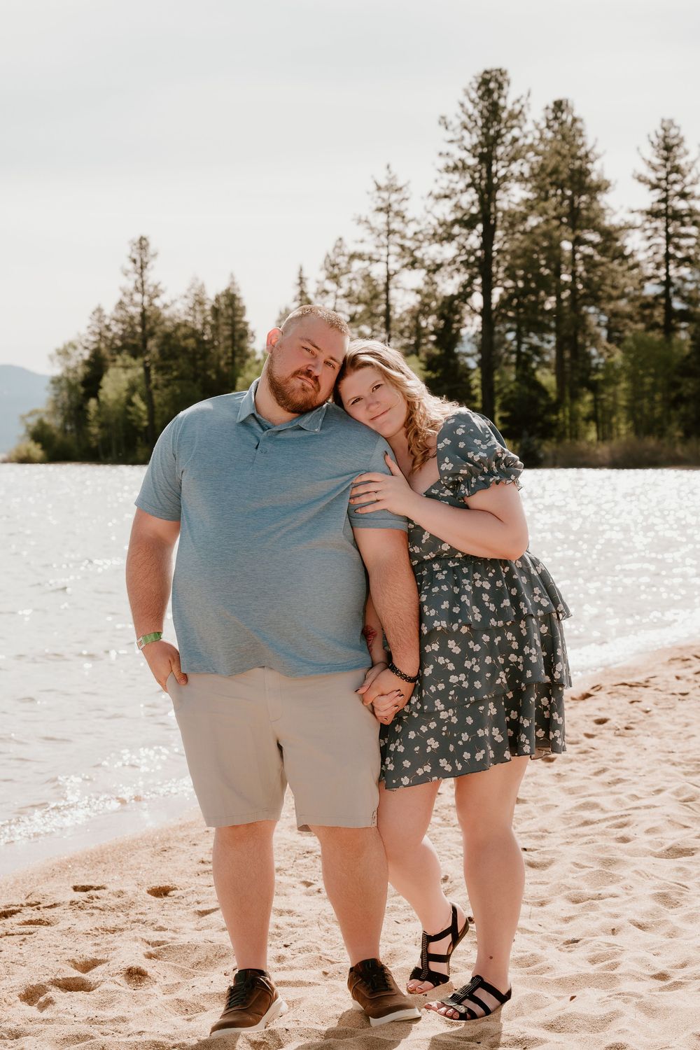 Couple embracing on sandy beach with pine trees and lake in background, woman in floral dress leaning on mans shoulder.