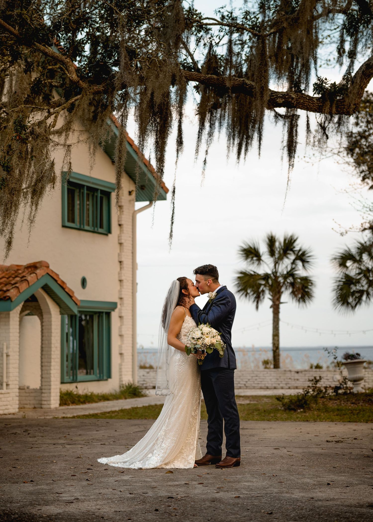 Bride and groom share a romantic kiss under Spanish moss-draped oak tree near waterfront venue with palm trees. Santa Rosa Beach Wedding