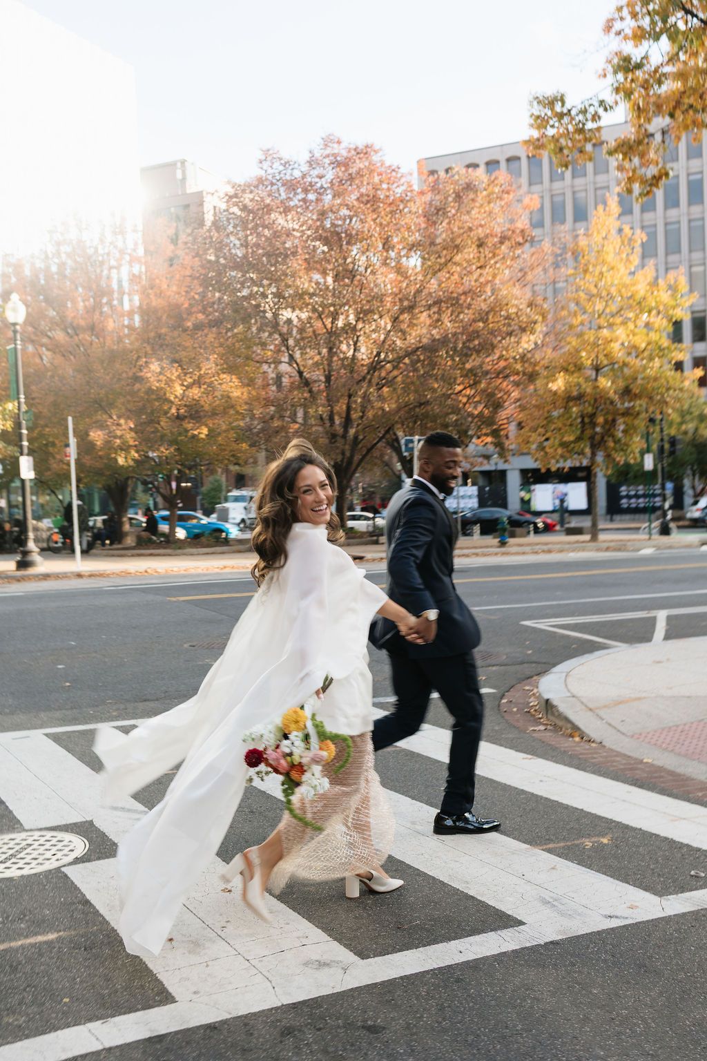 Bride and groom crossing a street in autumn, bride holding colorful bouquet with flowing white cape wedding dress.
