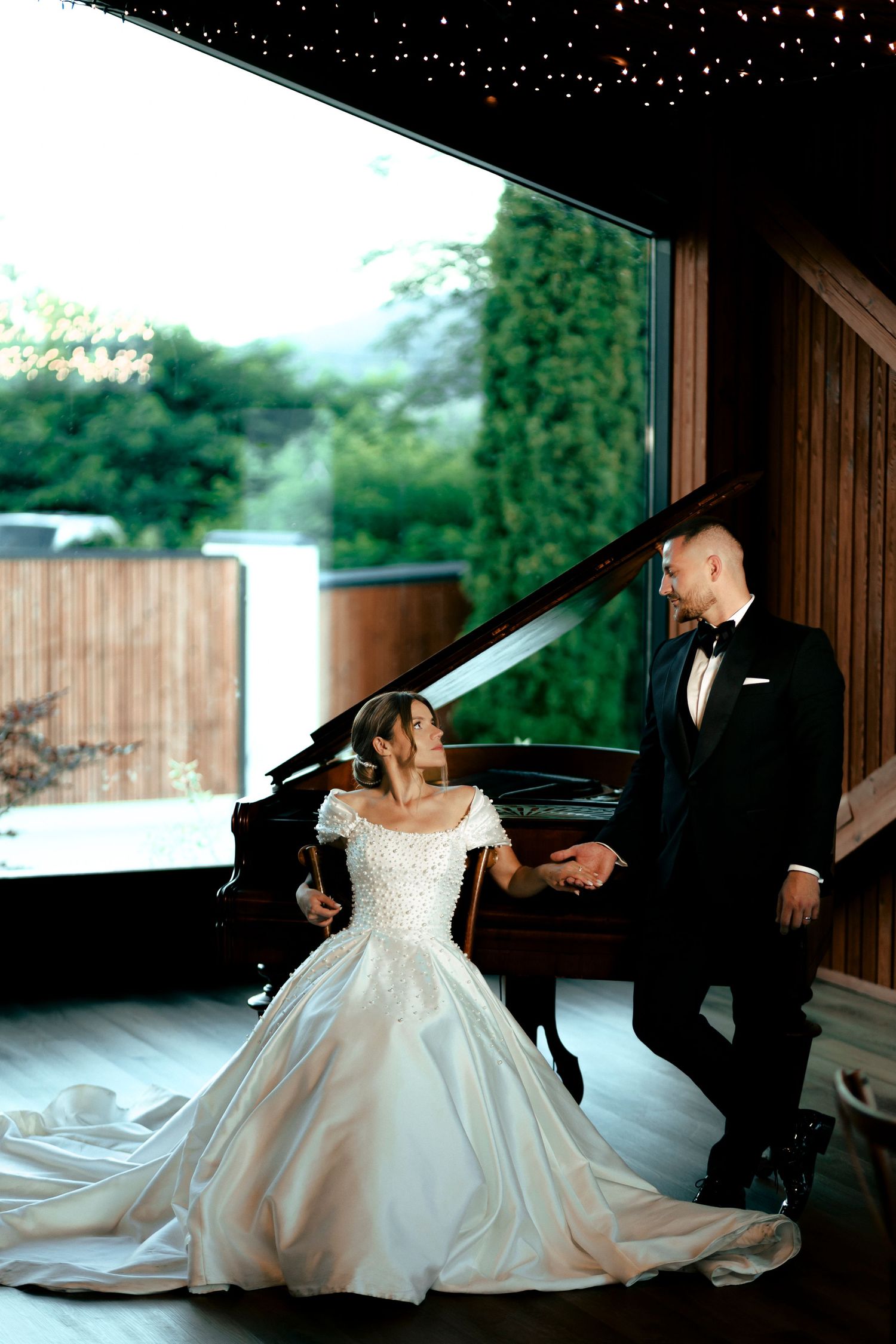 Bride in ball gown sitting at grand piano while groom leans in for a kiss.