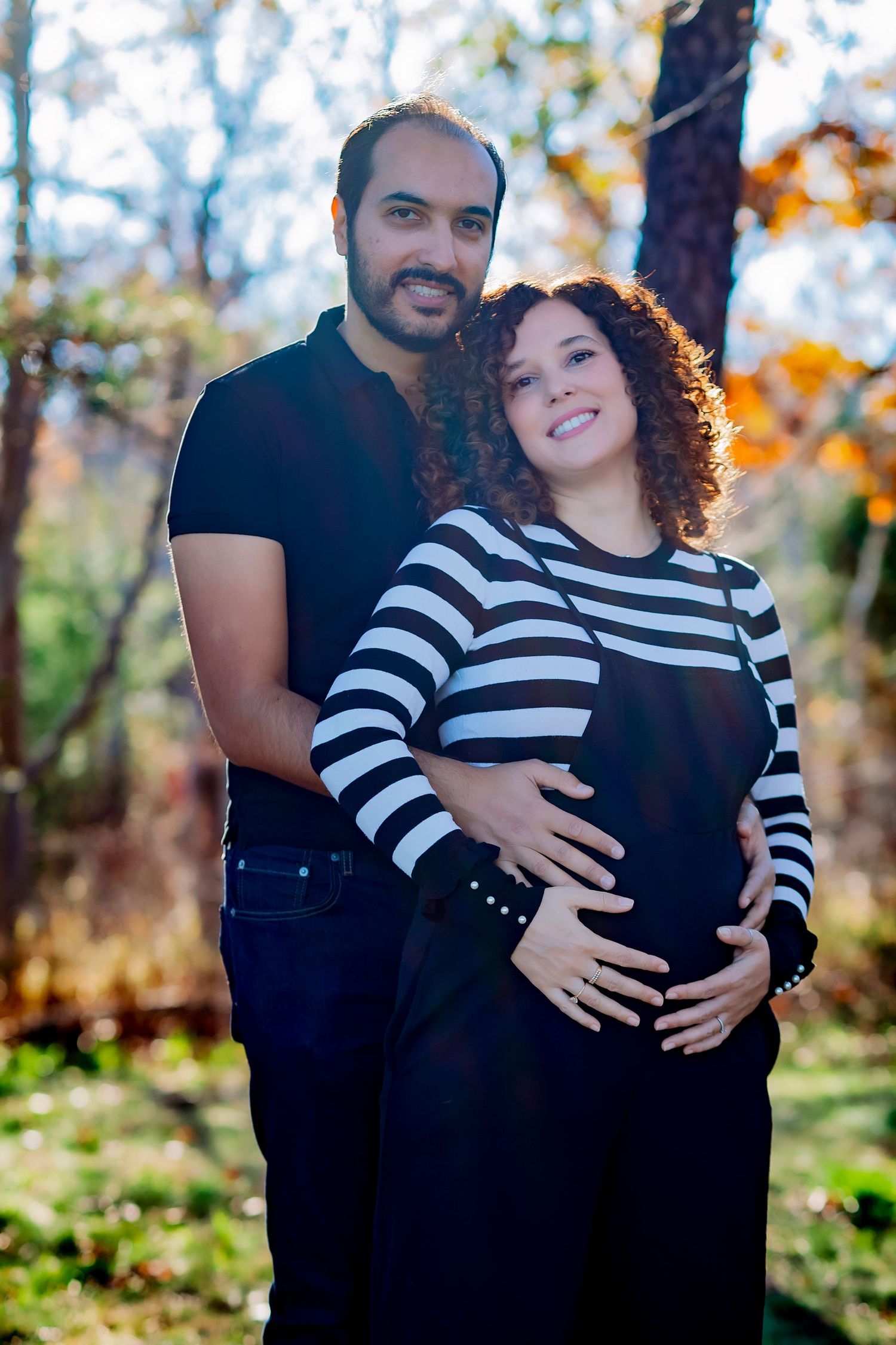 Happy expecting couple embracing outdoors in autumn, woman in striped top cradling baby bump.