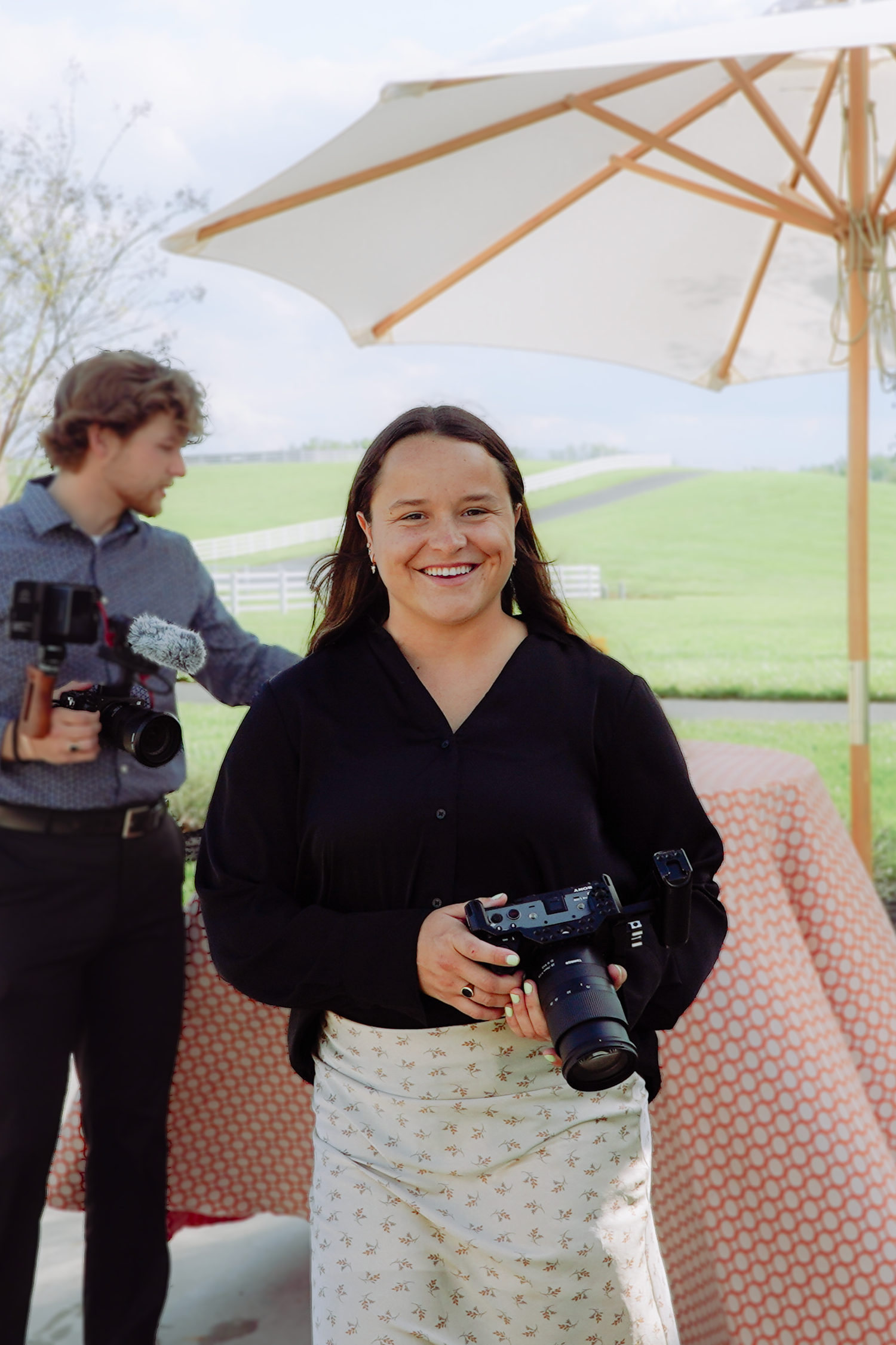 Smiling woman holding a DSLR camera outdoors with a white umbrella and green hills in the background.