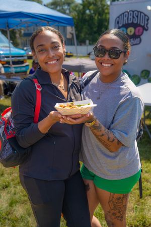 Two friends enjoying street food and smiling while holding paper plates outdoors at a local food event. Quad Cities Photography
