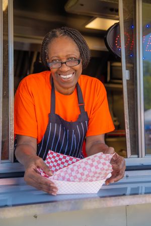 A food service worker in an orange shirt and striped apron smiles while folding paper in a restaurant service window Quad Cities Photography