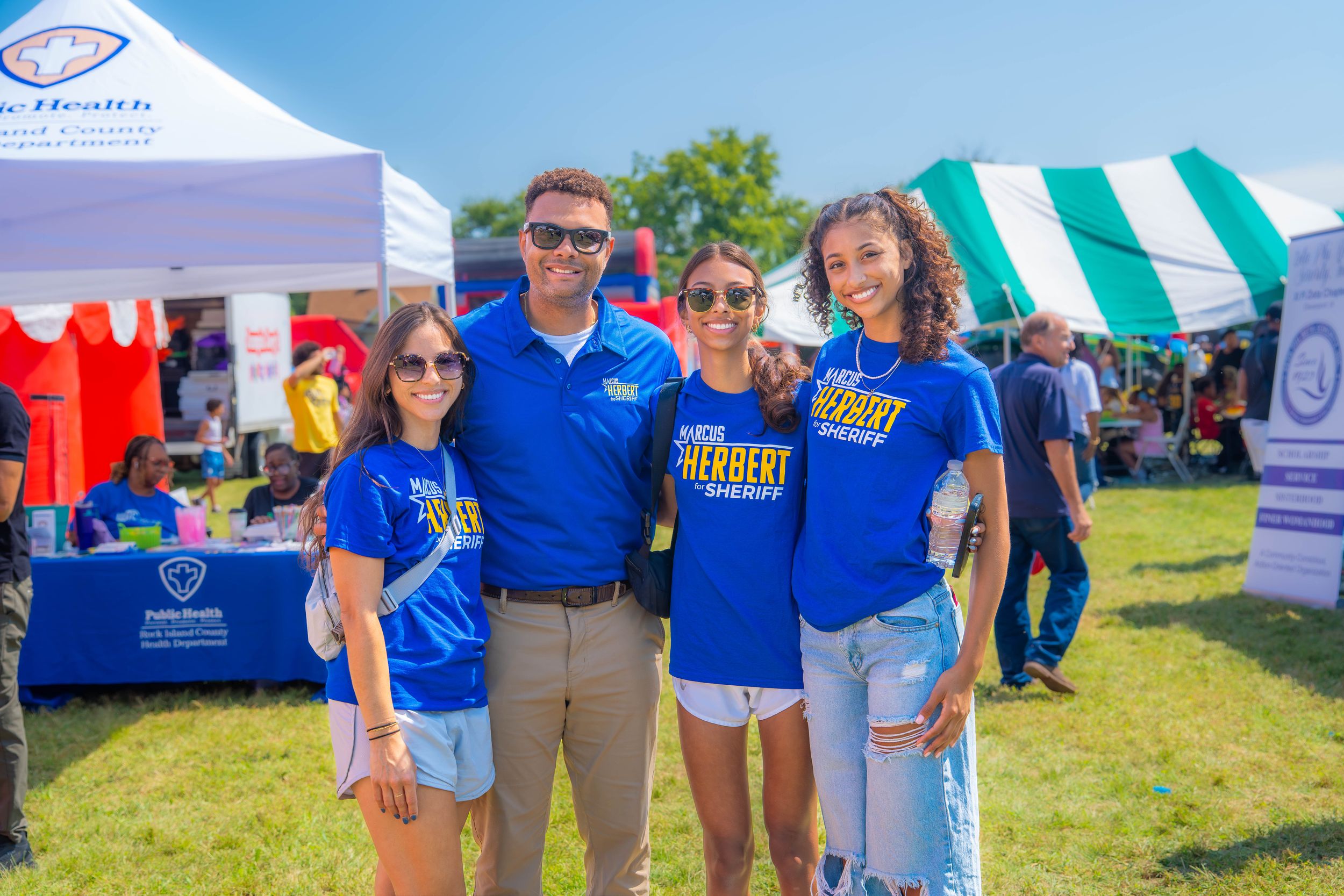 Group wearing bright blue volunteer t-shirts poses together at an outdoor community event with tents in background. Quad Cities Photography