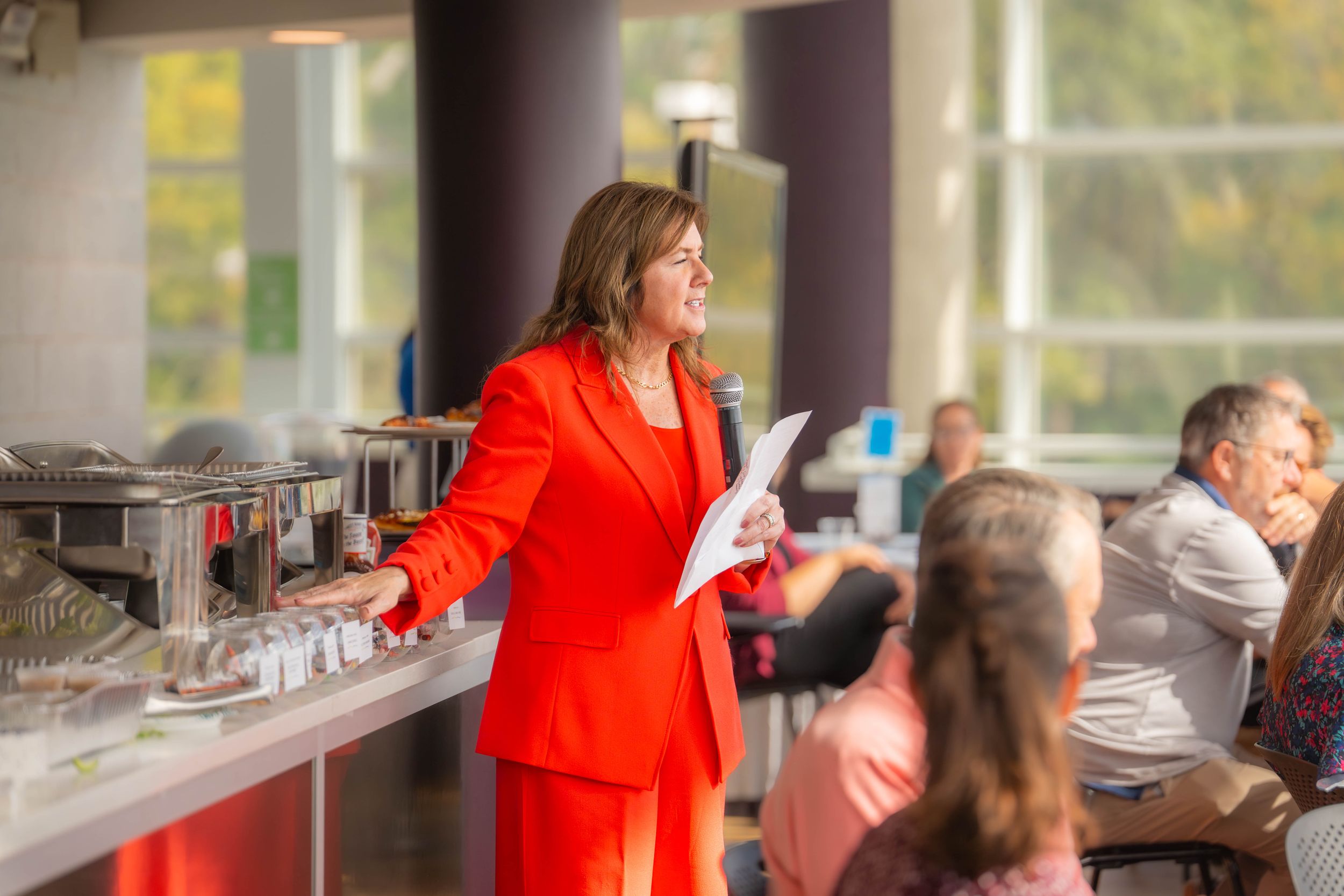 A person in a bright red blazer gestures while speaking to an audience in a conference room setting. Quad Cities Event Photography