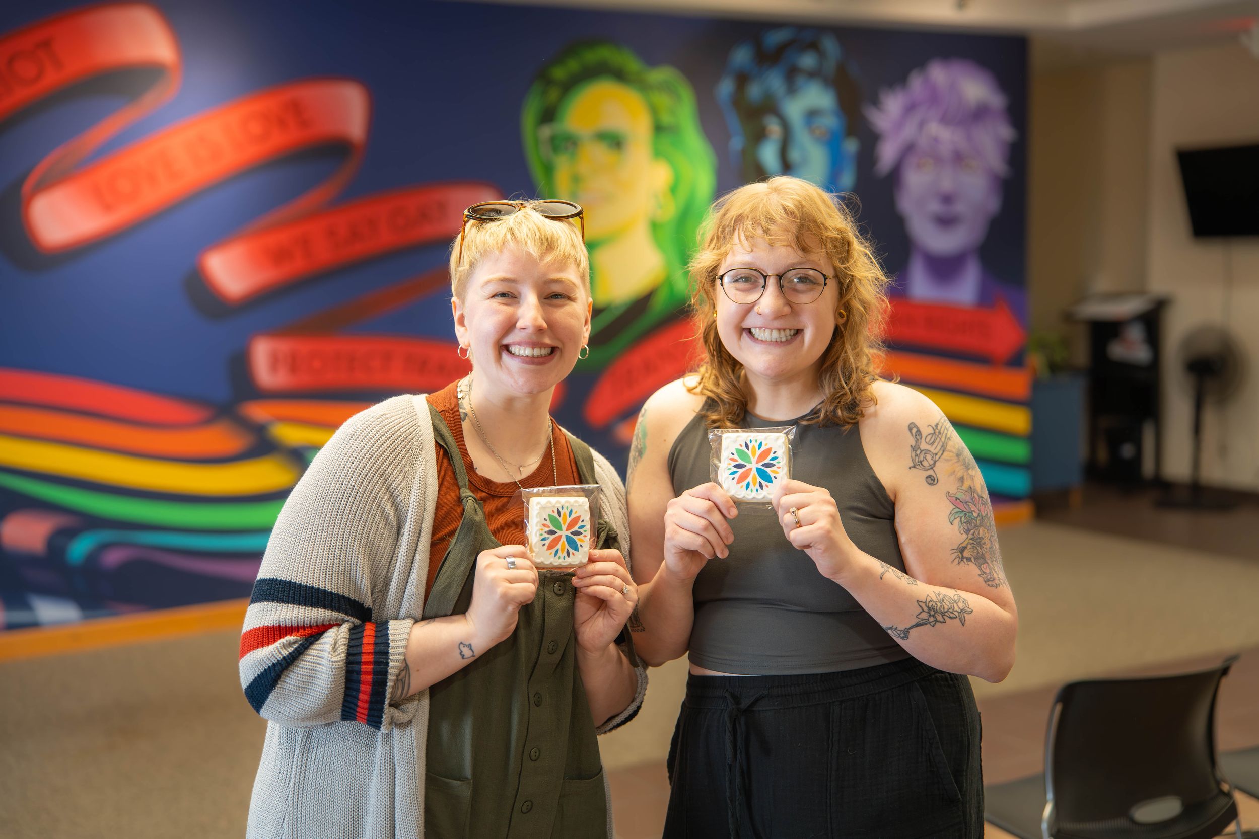 Two people smile while holding coffee cups in front of a colorful wall mural featuring rainbow stripes and artwork.
