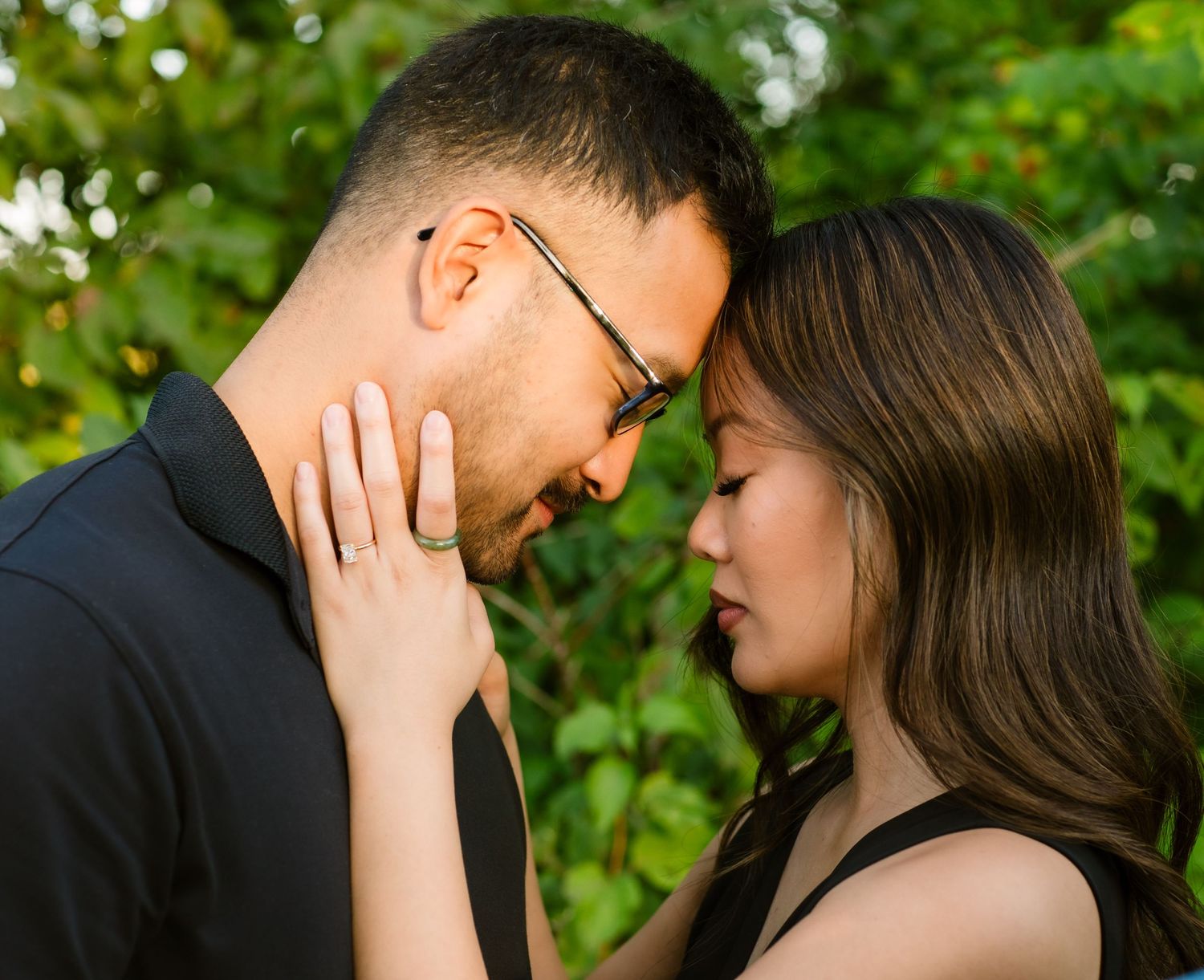 A tender romantic moment between a couple sharing a private embrace outdoors with green foliage in the background.