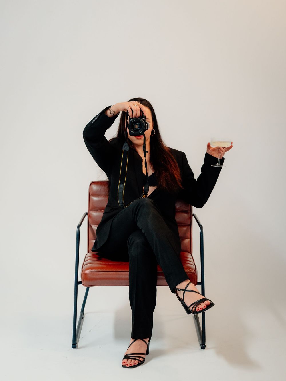 A photographer in black clothing sits on a brown leather chair taking photos against a white backdrop.