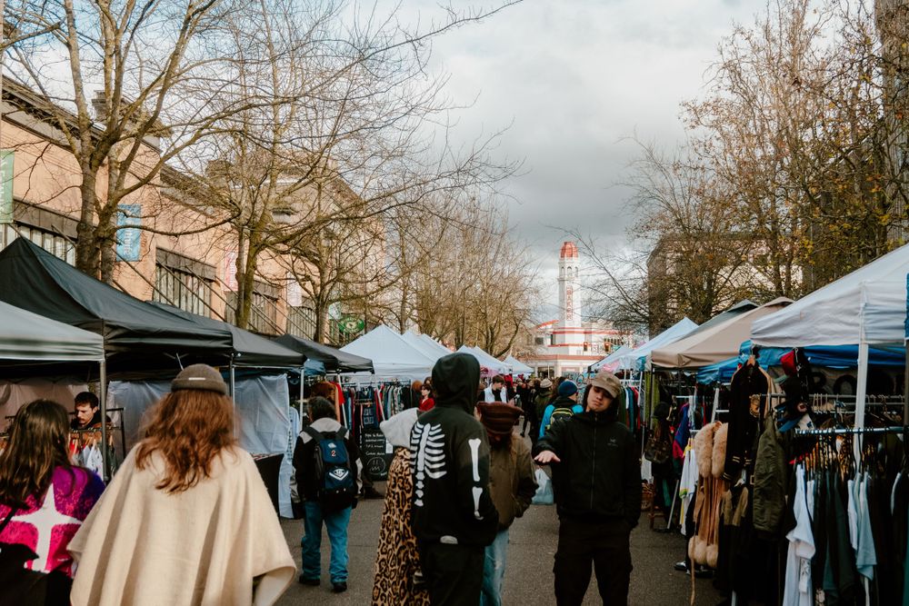 People shopping at outdoor market stalls under white canopies on cloudy day.