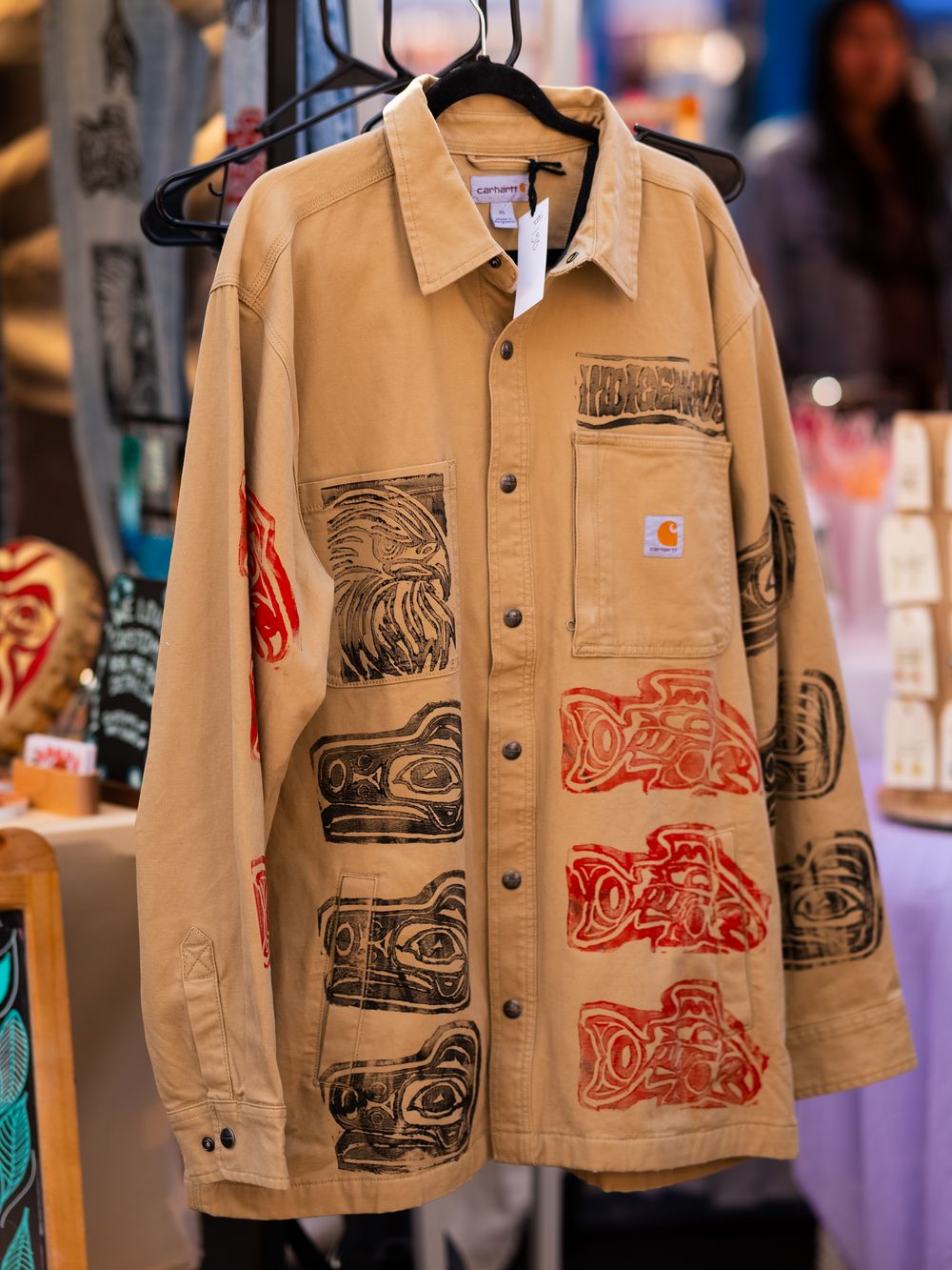 Tan canvas jackets with artistic block prints displayed on clothing racks at a market.