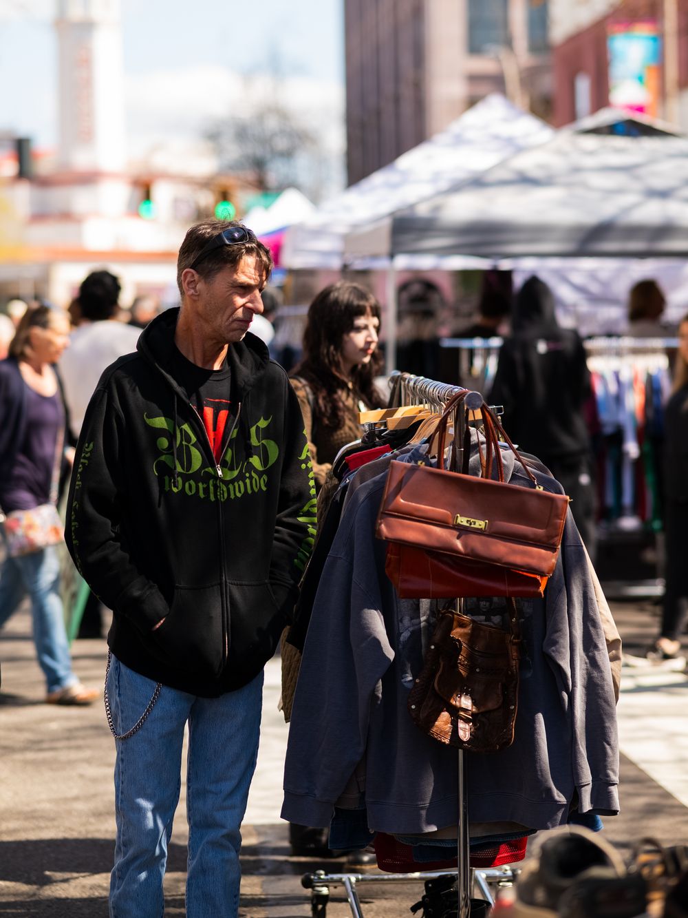 Shoppers examine merchandise at an outdoor market stall with tents in background.
