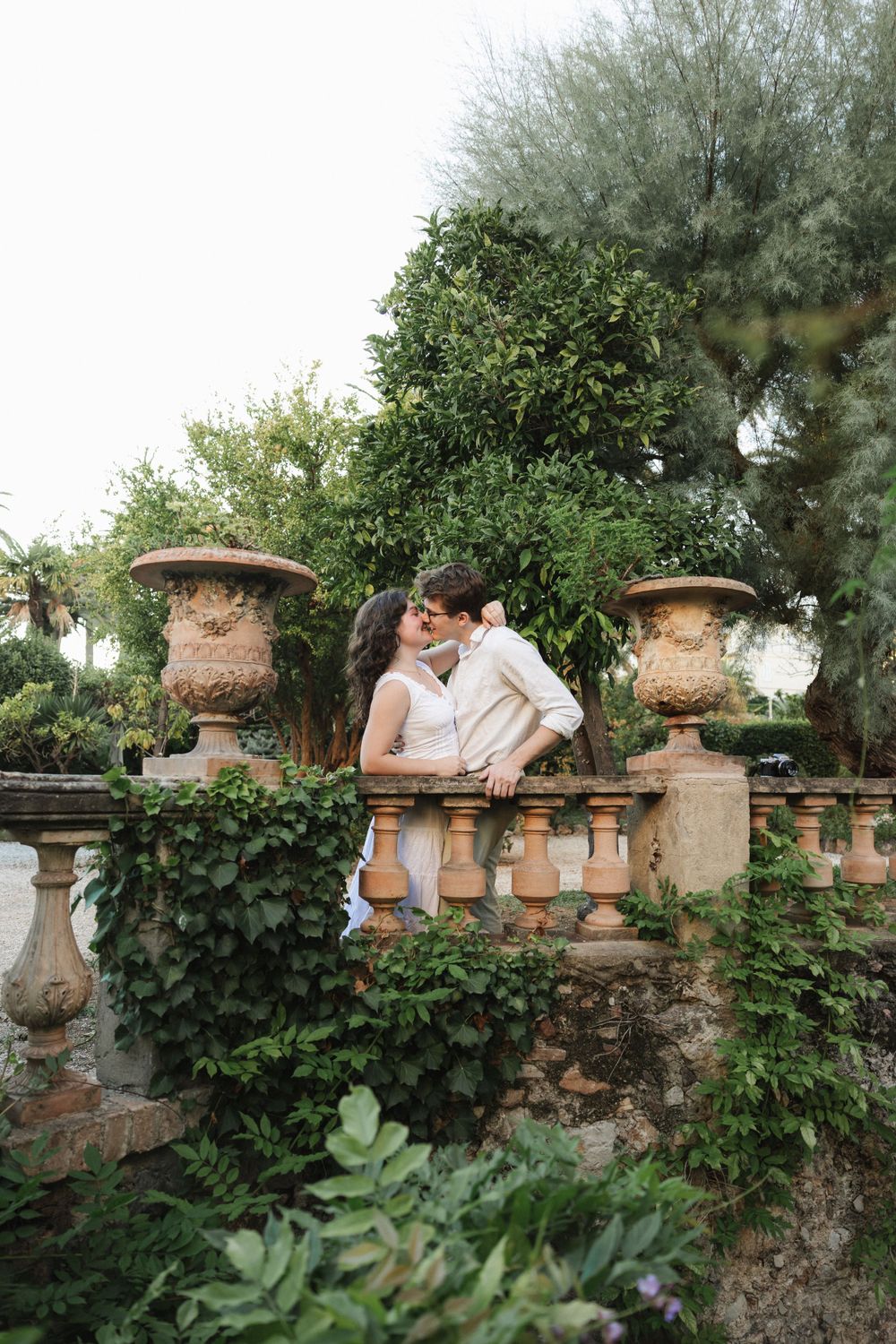 Couple sharing a kiss by an ornate stone balustrade surrounded by lush greenery.