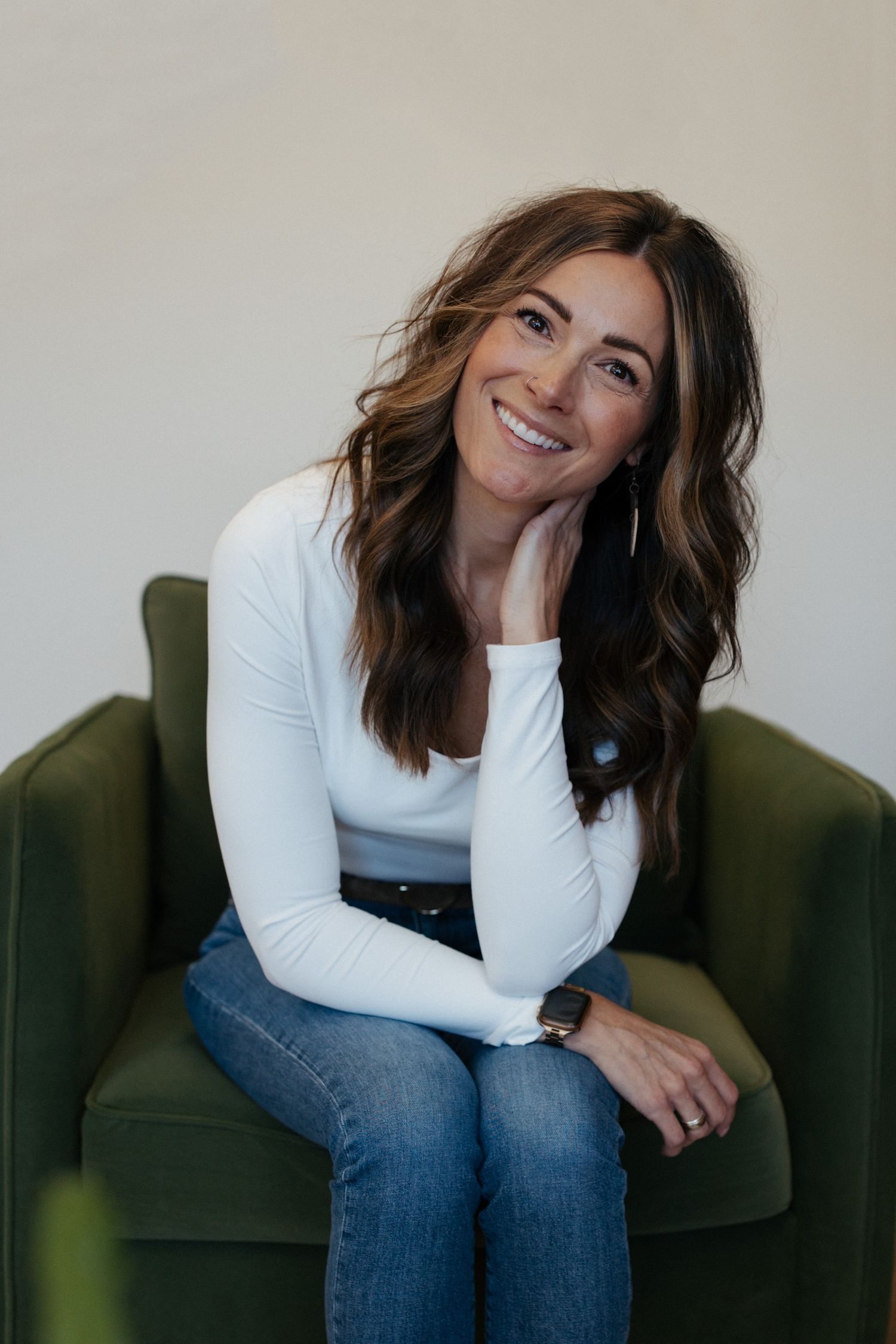 Woman in white long sleeve top and jeans sits smiling on green velvet chair.