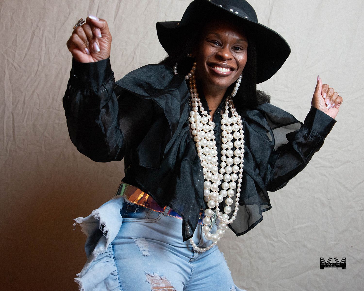 Energetic portrait of person in black hat and jacket with pearl necklaces and ripped jeans.