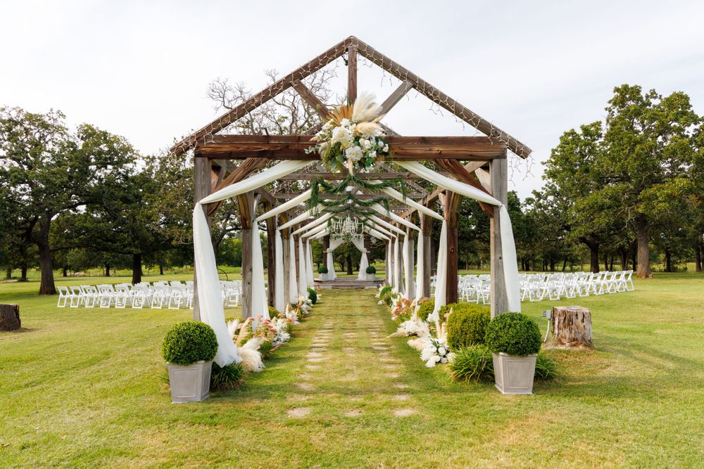 Rustic outdoor wedding ceremony setup with wooden arch and white chairs on grass.