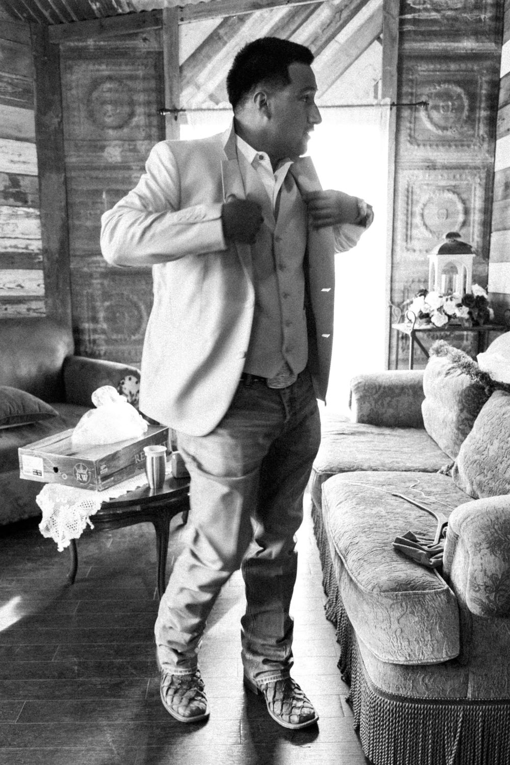Black and white photo of groom adjusting suit in rustic room.