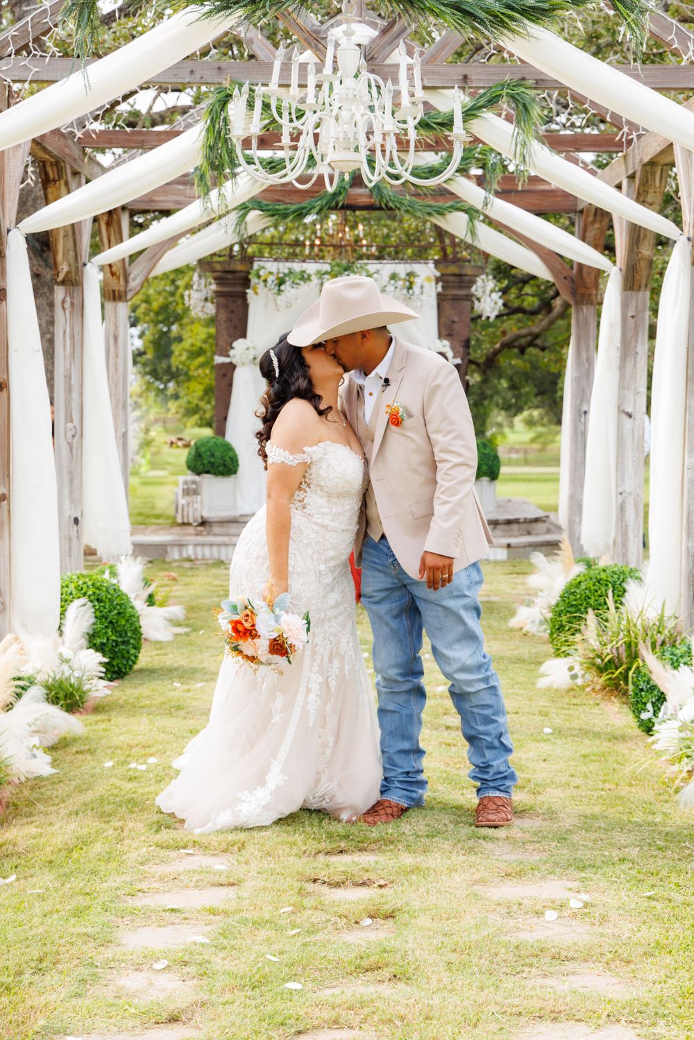Romantic moment under rustic wedding arch decorated with white fabric, greenery and chandelier.