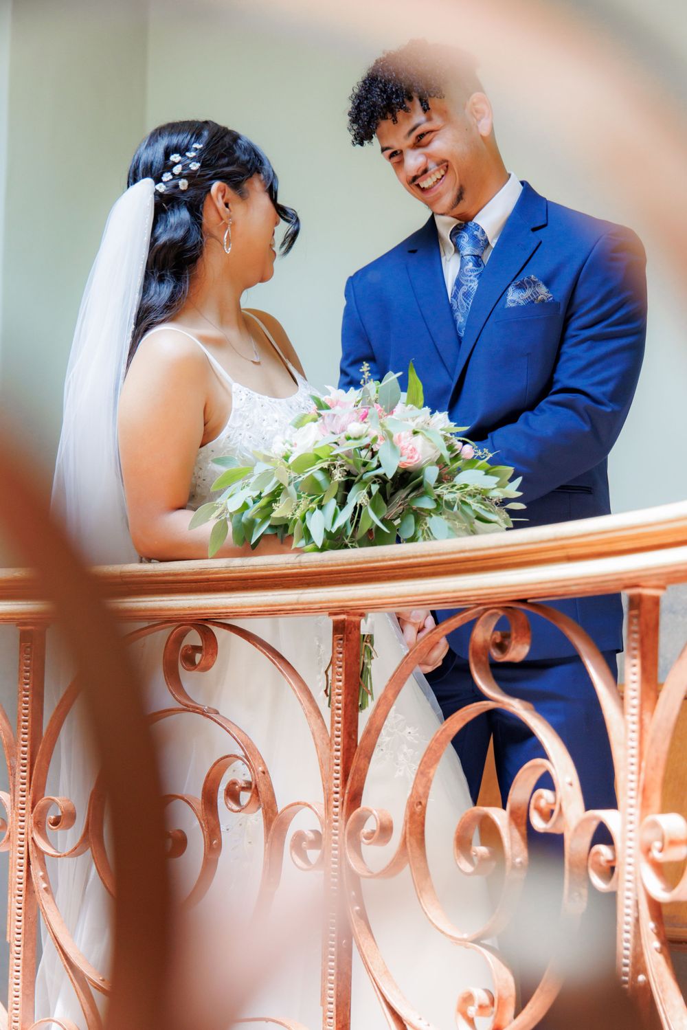 A joyful wedding couple sharing a laugh on ornate stairs while holding a bouquet of flowers.