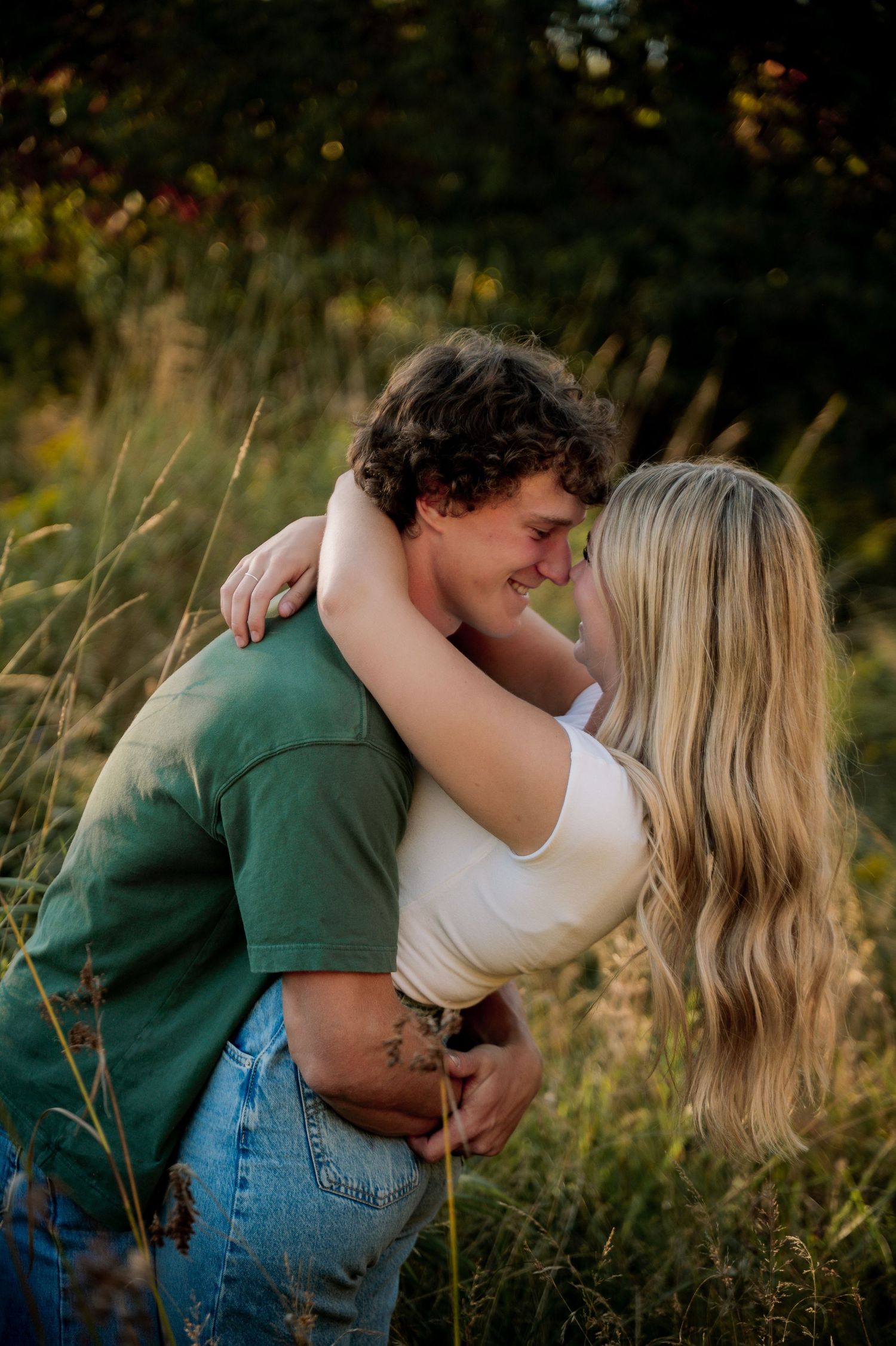 Couple embraces by tranquil lakeside under leafy green trees.