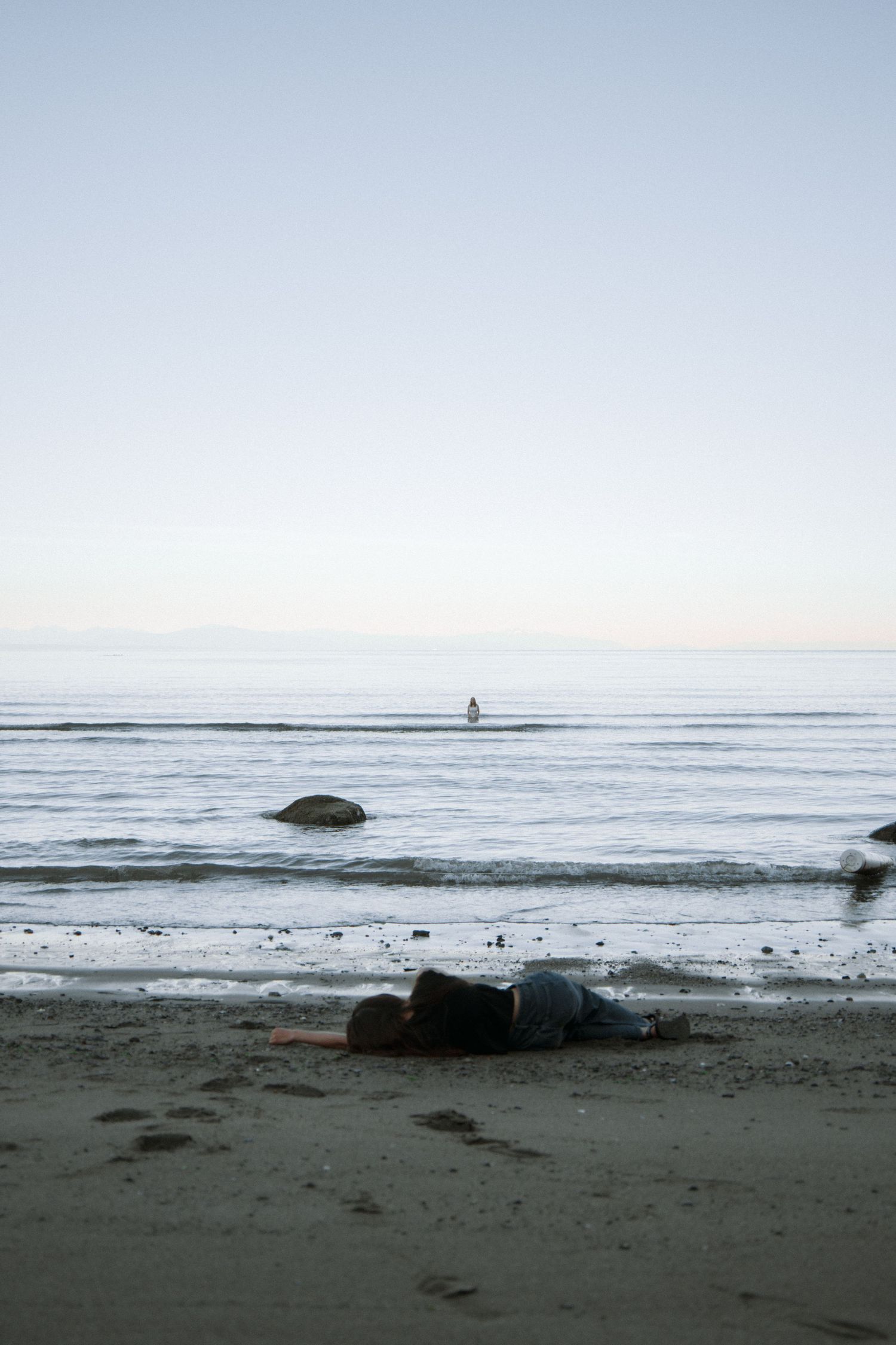 A solitary figure lays on a sandy beach at dusk with a distant silhouette visible on the calm ocean horizon.