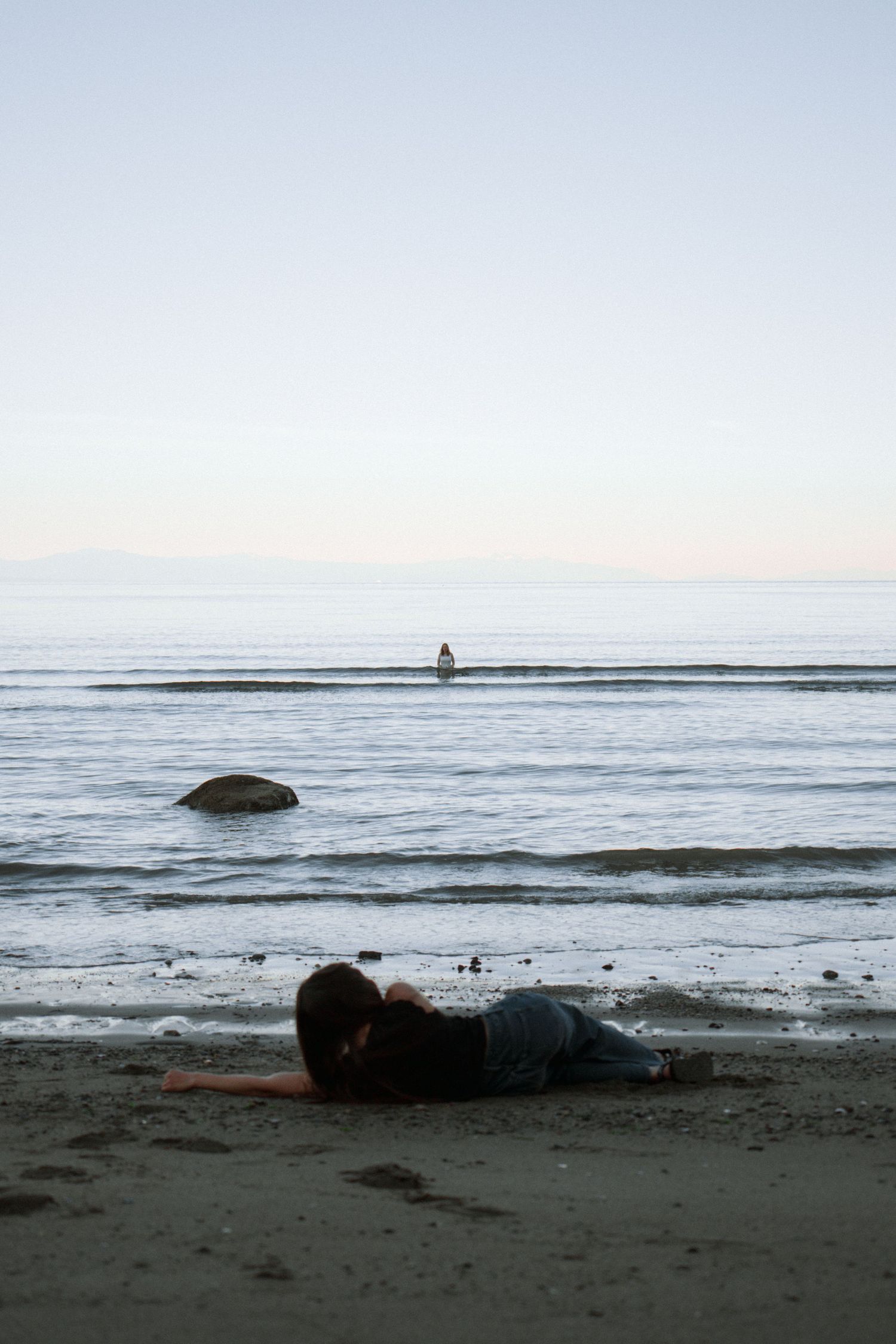 Figure resting on beach sand during sunset with gentle waves and a lone silhouette in the distance.