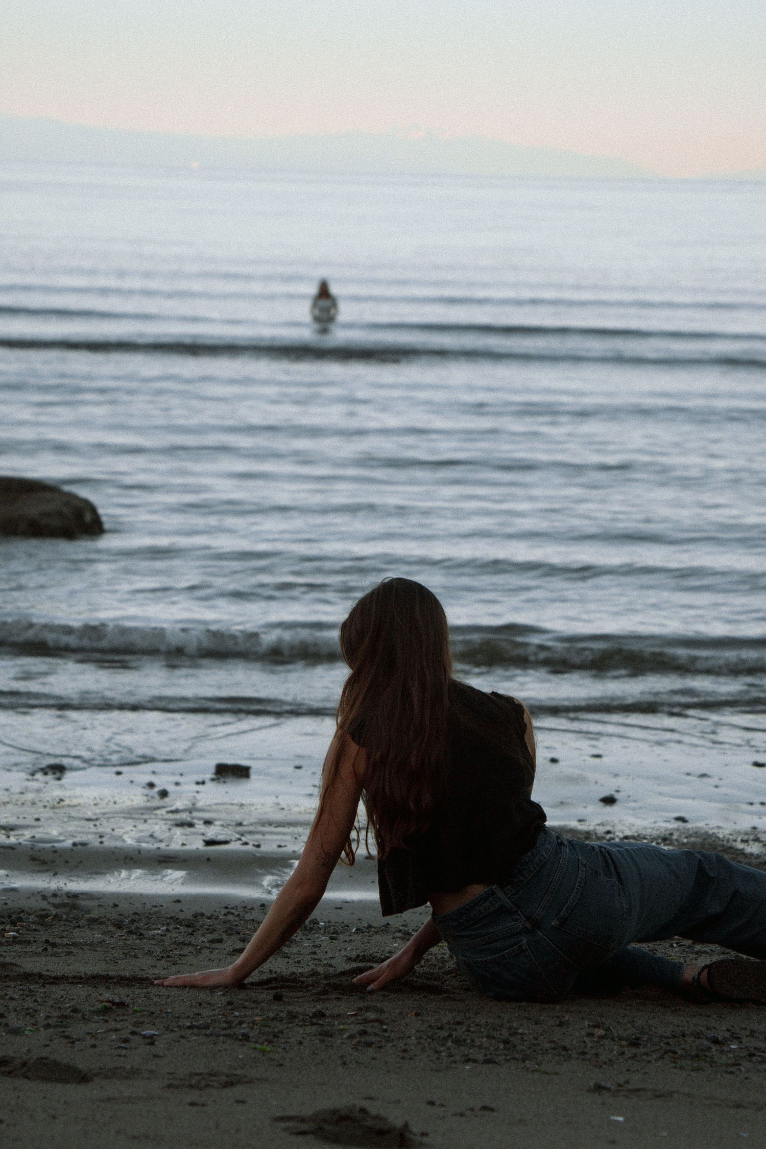 Person lounging on beach at twilight watching distant figure in ocean waters.