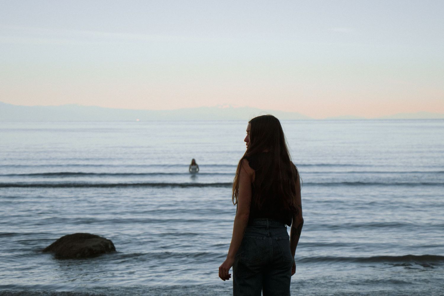 Person in dark clothing gazes out at lone figure standing in shallow ocean waters during sunset.