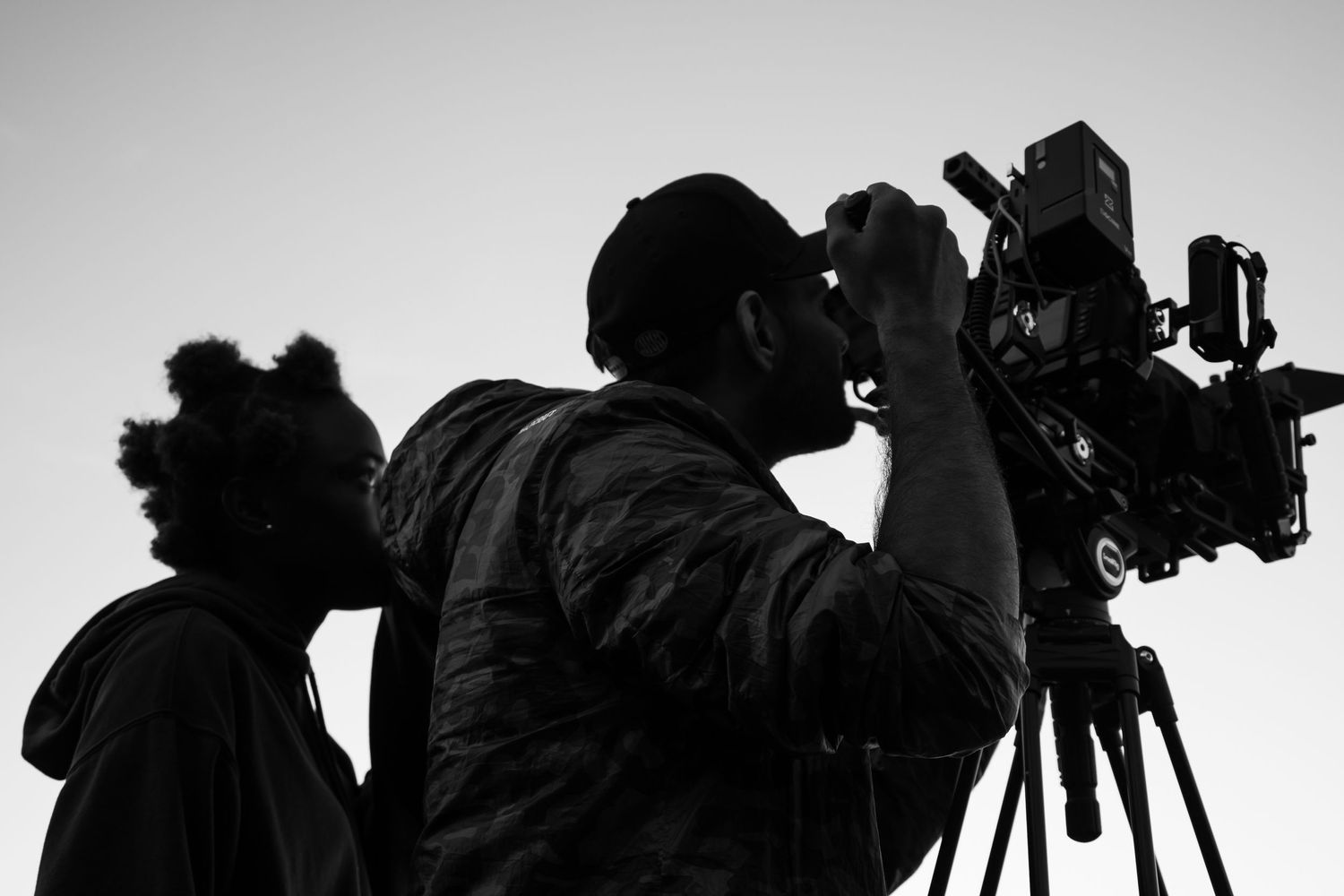 Silhouette of two people operating a professional film camera against a light background.