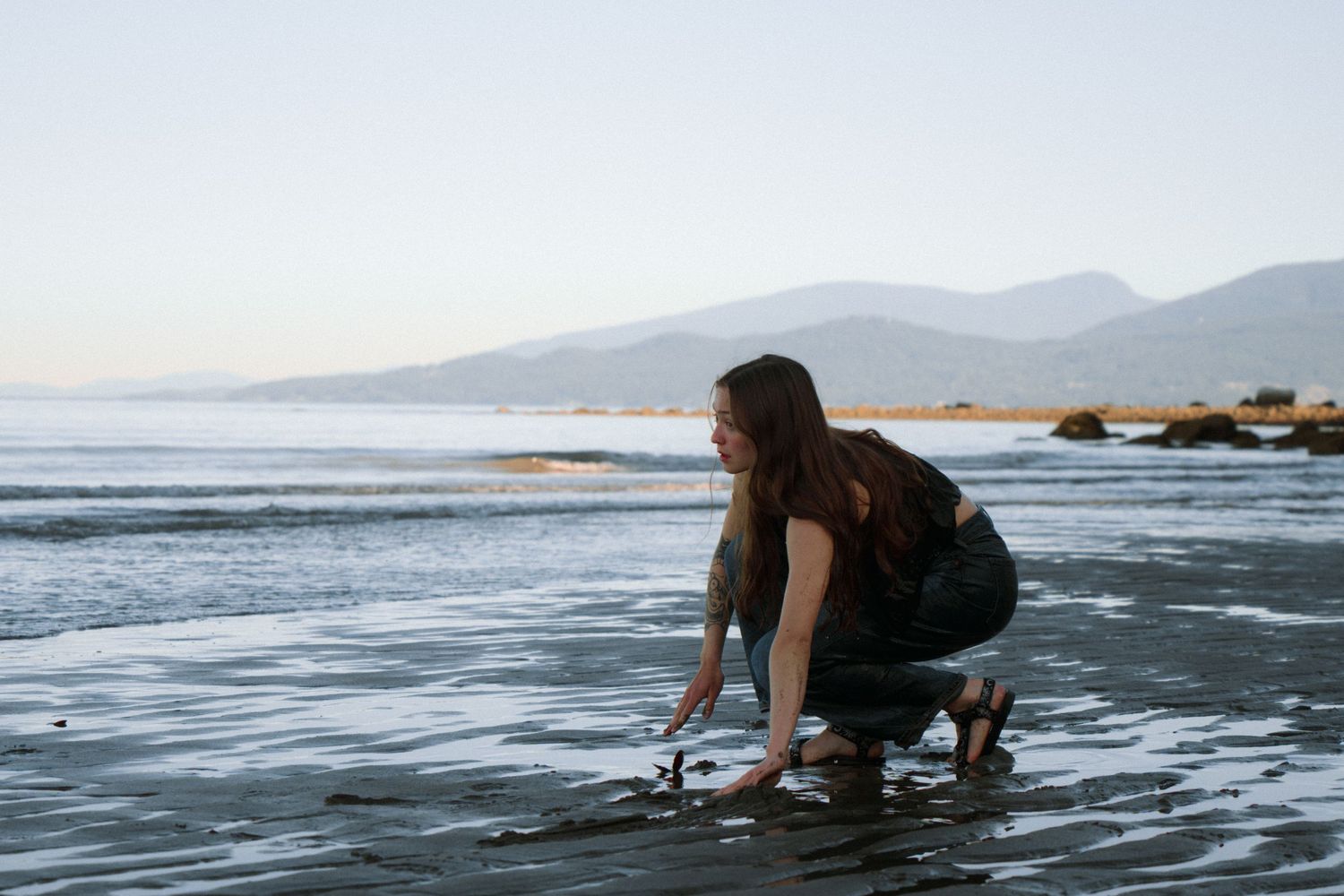 Person crouching near ocean shore with mountains visible in background during golden hour.