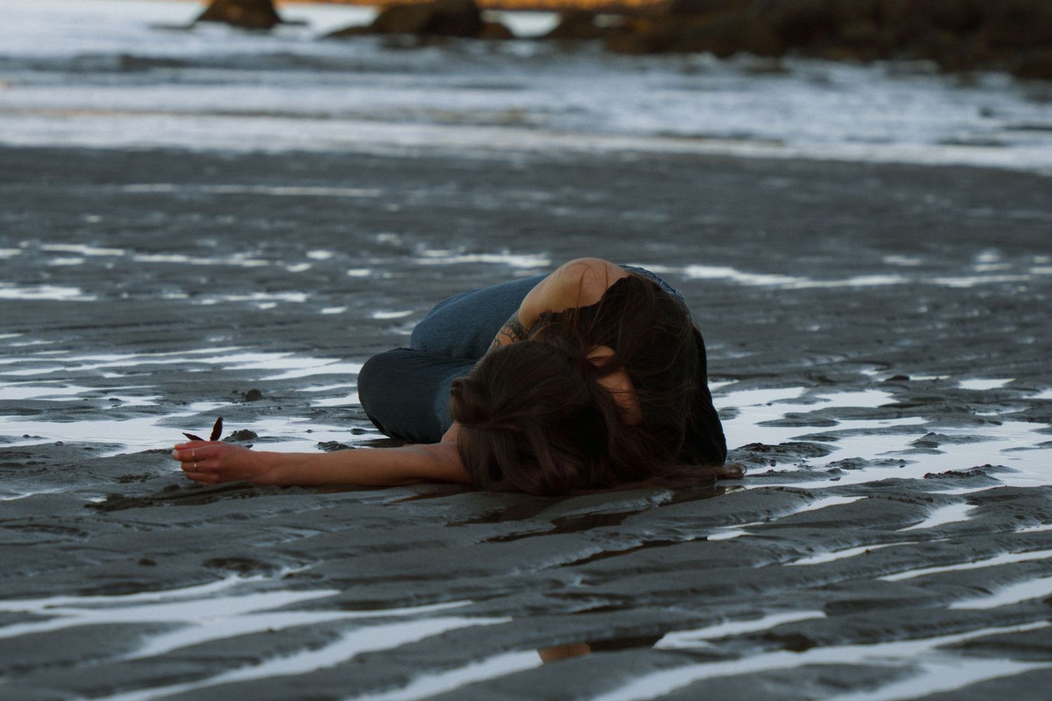 Figure lying on wet sand near ocean shore during moody twilight.
