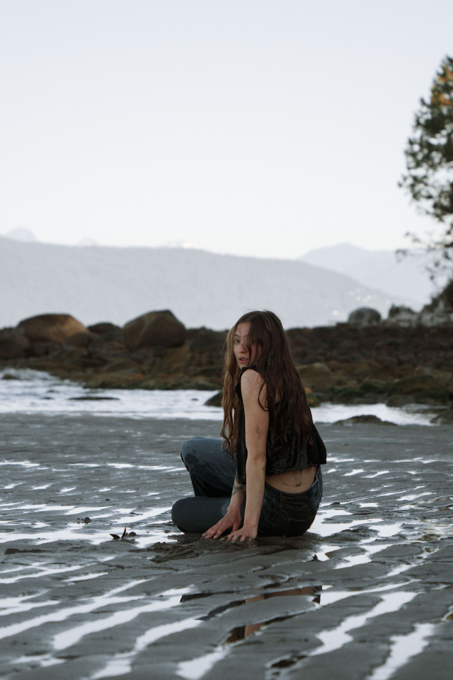 Person sitting on rocky beach with mountains and evergreen trees in background at dusk.