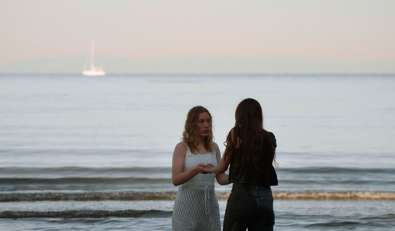Two people standing together at sunset on a beach with a sailboat visible in the distance.